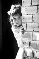 Black and white image of two year old little girl peaking around brick wall